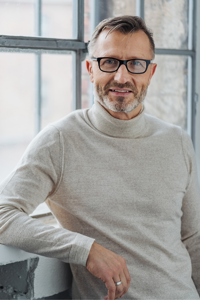 Smiling attractive mature man wearing glasses leaning on the sill of a large bright window with copy space looking at the camera